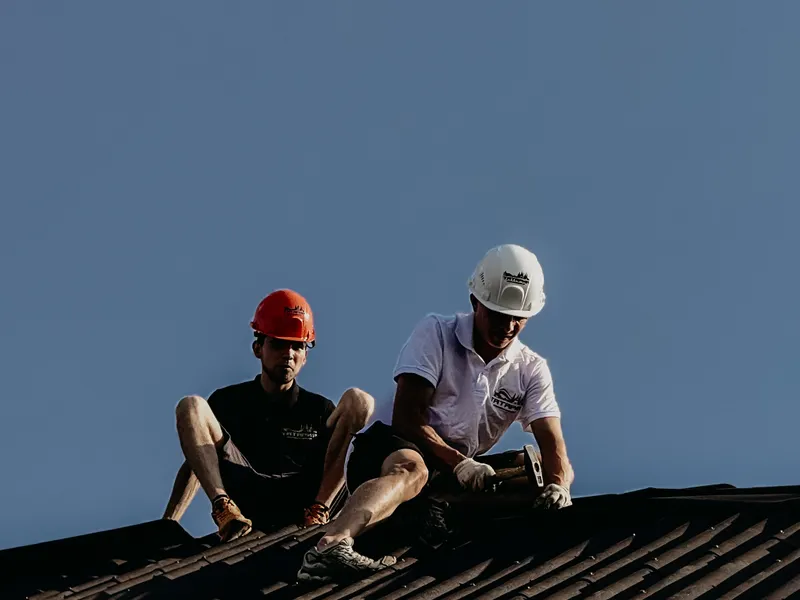 Two workers in hard hats seated on a dark roof slope against a clear sky