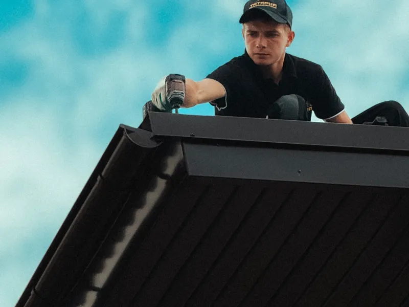 Worker fastening trim at the eave of a dark metal roof, drill in hand against a blue sky