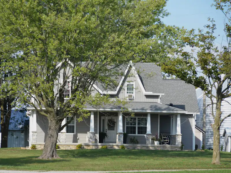 Gray craftsman-style home with gabled shingle roof and covered porch behind a broad shade tree