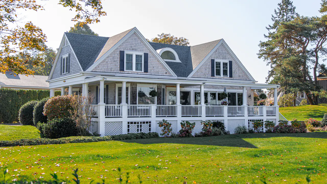 Gray two-story farmhouse with wraparound porch and architectural asphalt shingle roof, autumn trees behind
