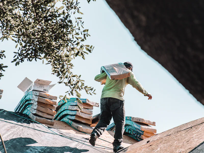 Worker carrying a shingle bundle across a roof with stacked material staged behind