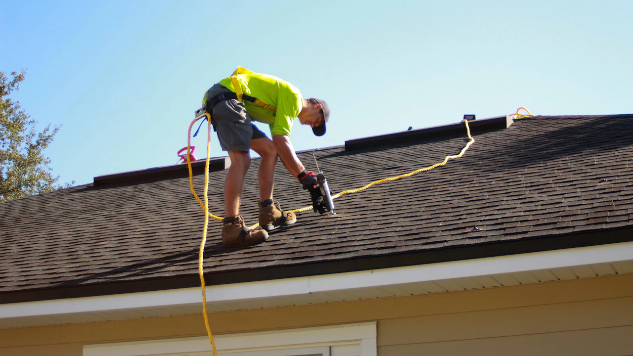 Fastening architectural shingles on a roof slope against a clear sky — representative