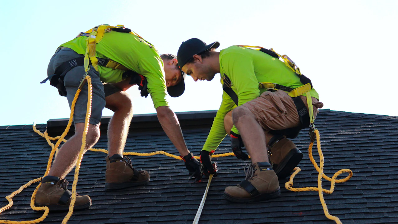 Harnessed roofers working architectural shingles near the ridge, safety ropes set — representative