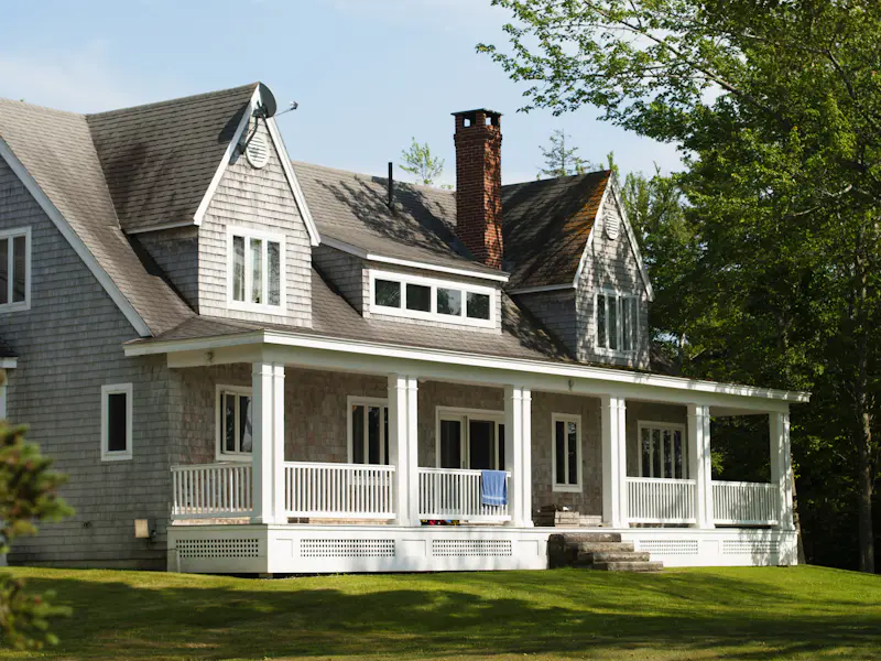 Shake-sided cape with brick chimney, dormers, and gray shingle roof behind a deep front porch