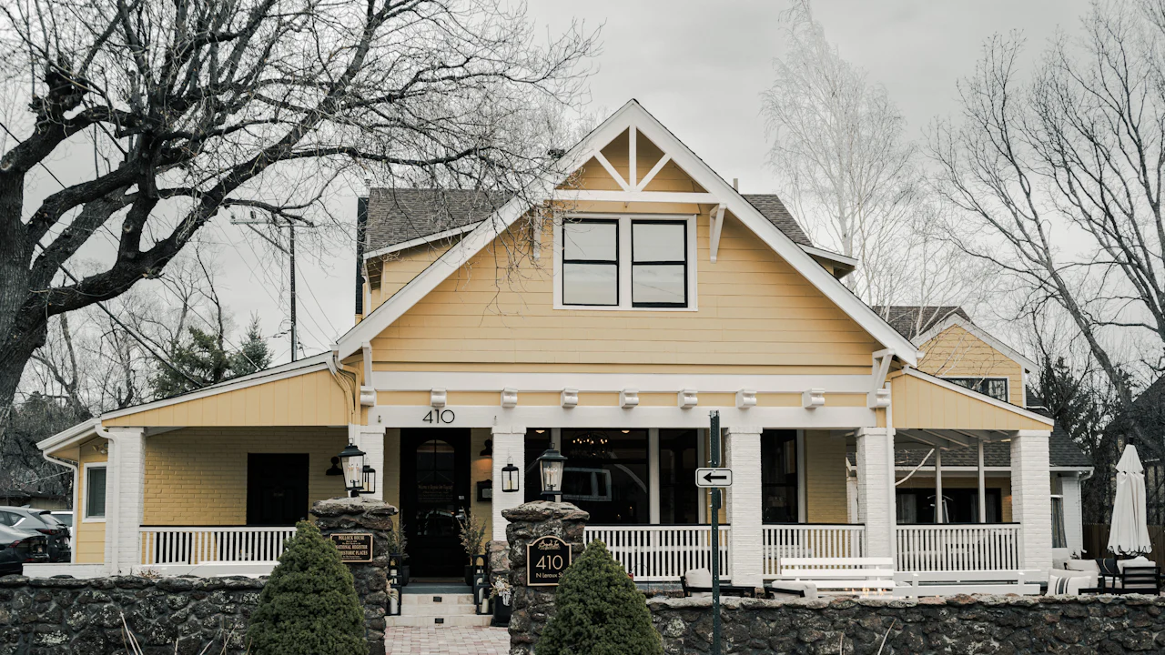 Yellow craftsman home with wide gable roof and stone wall under a gray winter sky, bare trees